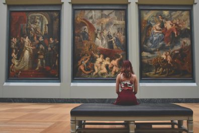 Une jeune fille est assise de dos dans un musée. Elle est face à trois grands tableaux.